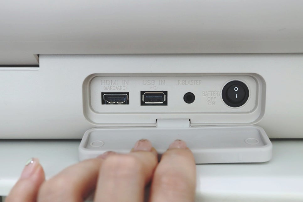 스탠바이미 Go 후면 포트 덮개를 손으로 열고 있음. 내부에는 HDMI 입력, USB 입력, IR 블라스터, 배터리 스위치, 전원 버튼 있음.